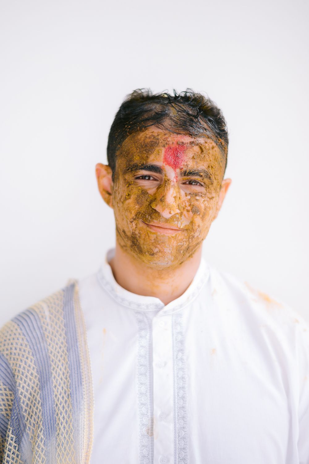 Man with turmeric paste and red tilak on forehead smiling, wearing white kurta during Haldi ceremony.