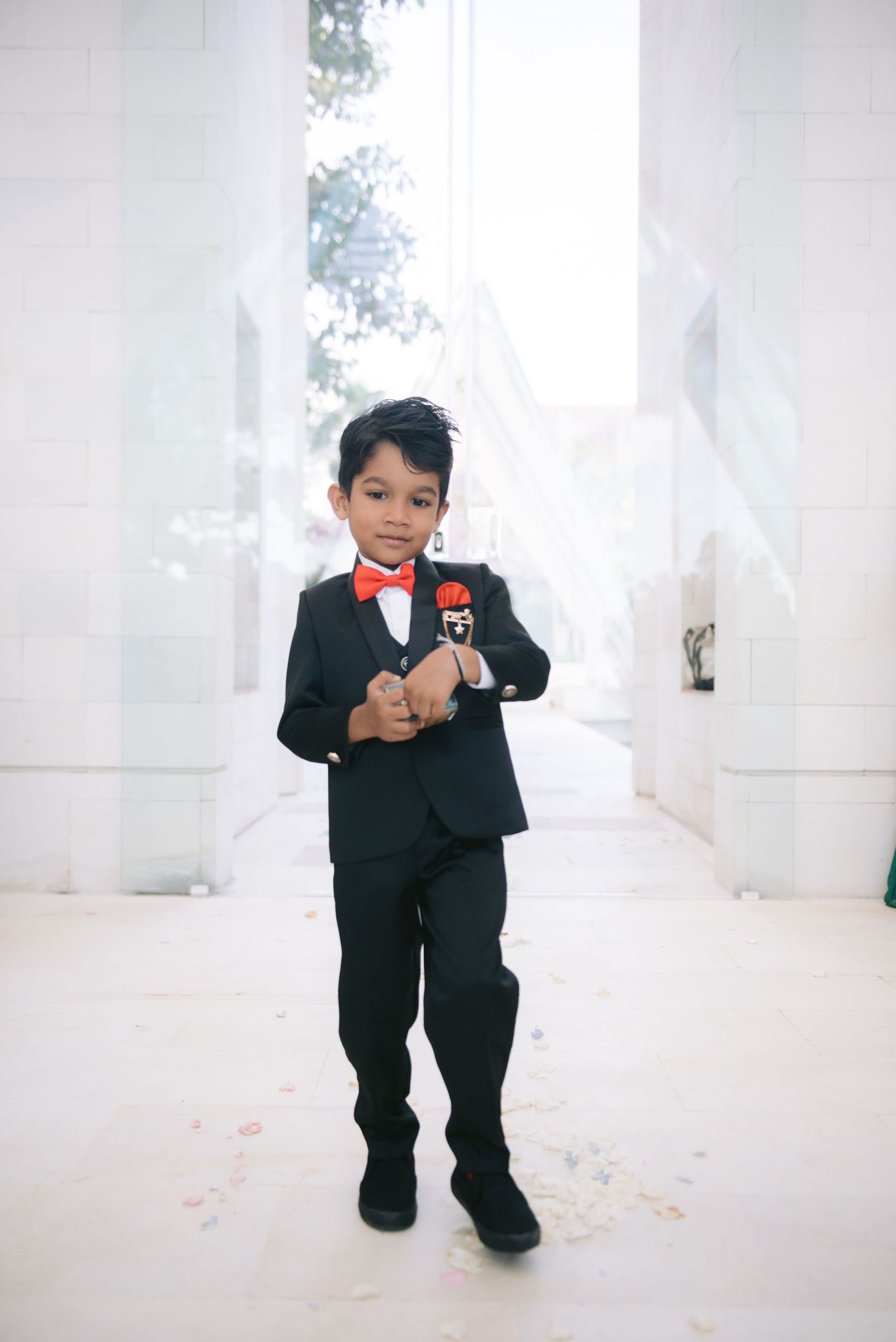 Young boy in black tuxedo with red bow tie holding a microphone at a wedding ceremony.