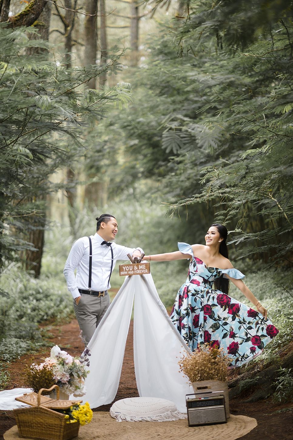 Couple laughing by a white teepee tent in a forest, woman in floral dress, man in suspenders, romantic outdoor picnic setup.