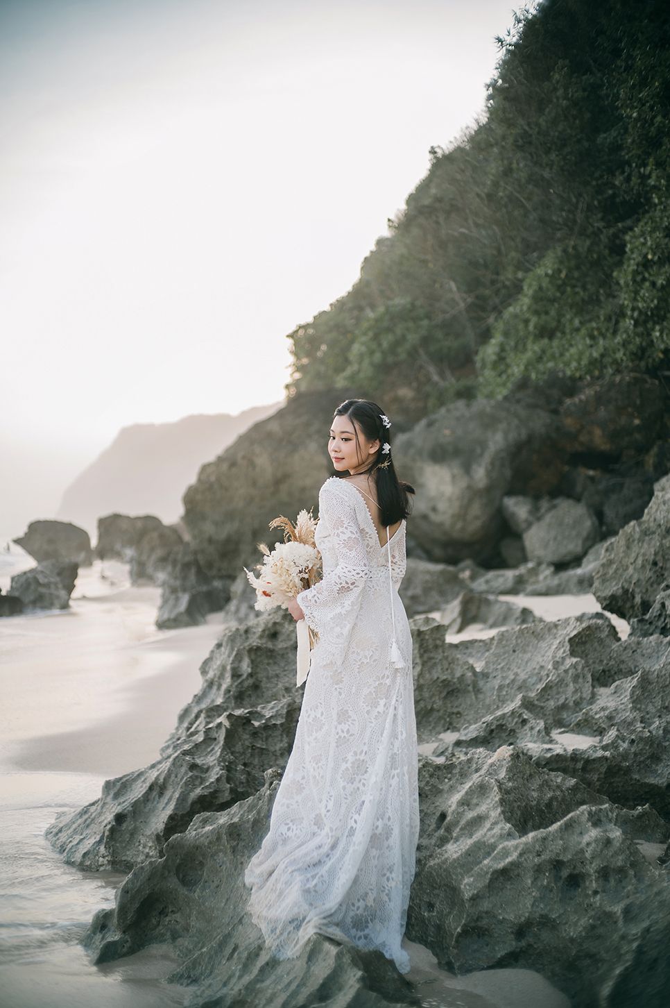Bride in white lace gown holding bouquet on rocky beach with green cliffs and ocean backdrop.