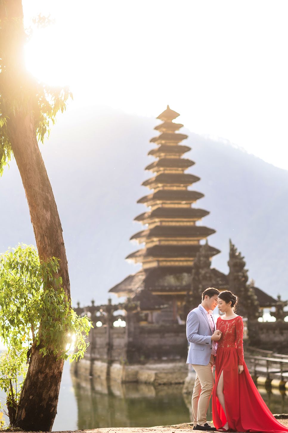 Couple in red dress poses romantically before Pura Ulun Danu Bratan temple in Bali with misty mountains behind.
