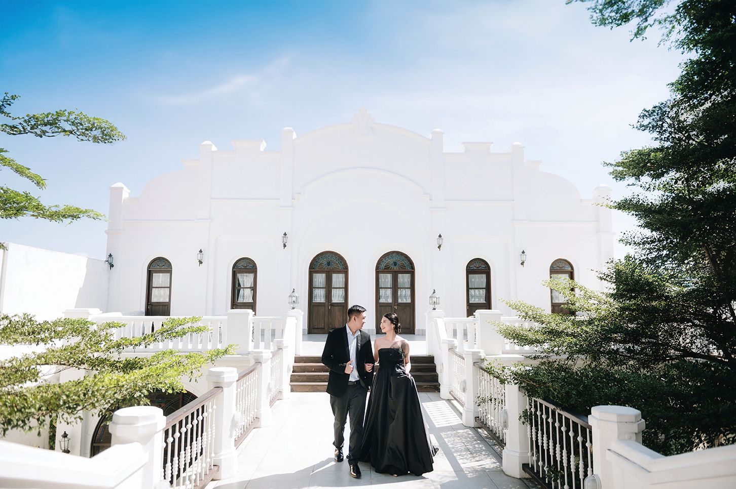 Couple in black formal wear poses on white steps before elegant white building with arched windows and lush trees.