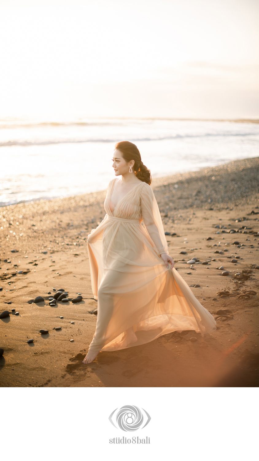 A woman in a flowing peach gown walks gracefully on a sandy beach at golden sunset hour.