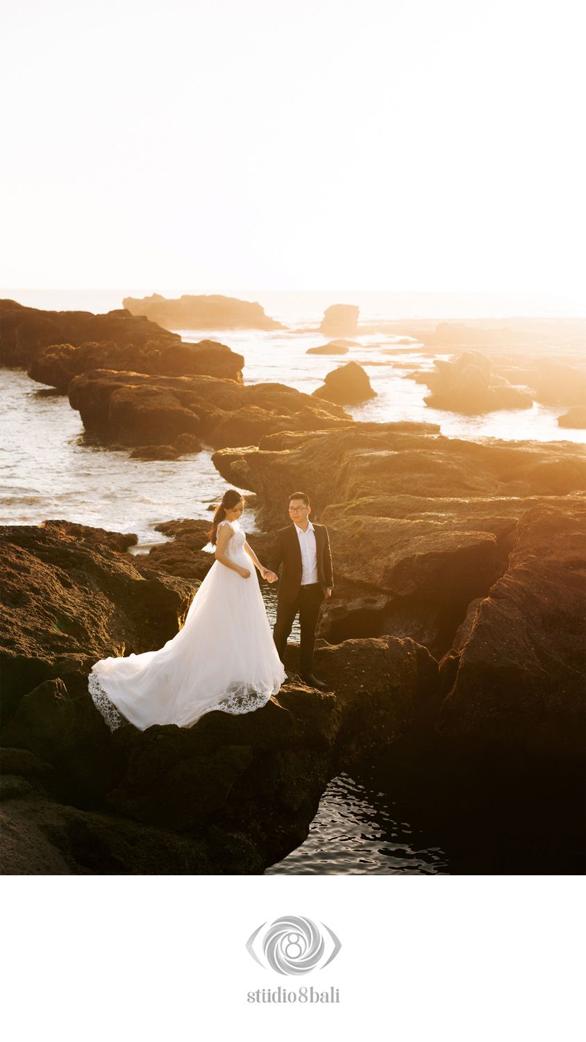 Bride and groom standing on rocky coastal cliffs at golden sunset during Bali pre-wedding photoshoot.