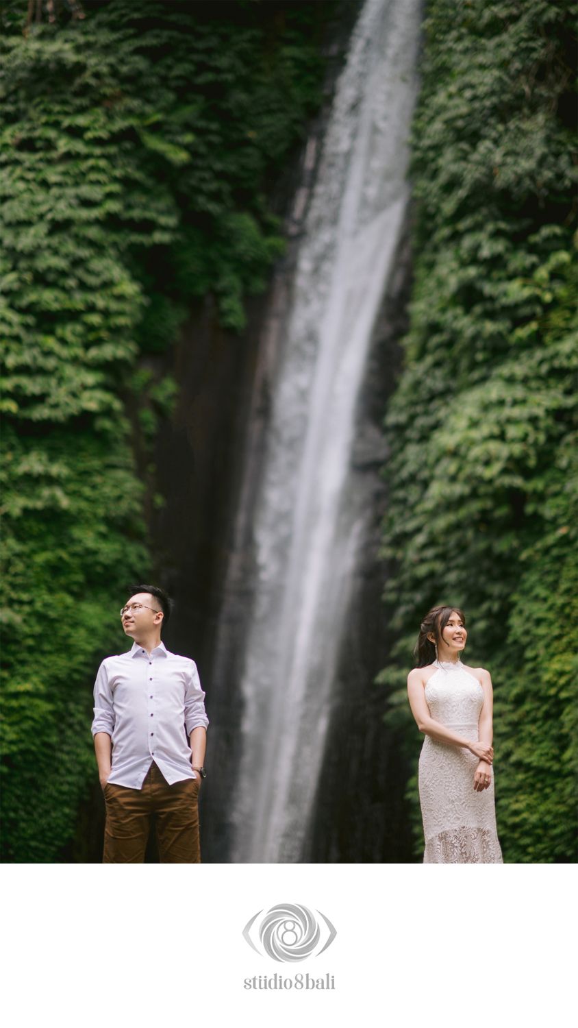 Couple poses before a tall waterfall surrounded by lush greenery in Bali, man in white shirt and woman in lace dress.