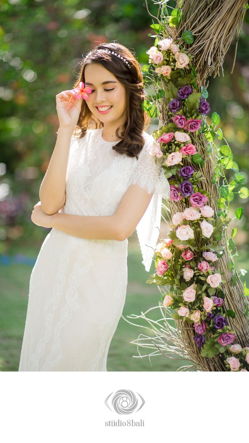 Smiling bride in white lace dress poses beside a floral arch with pink and purple roses in a garden setting.
