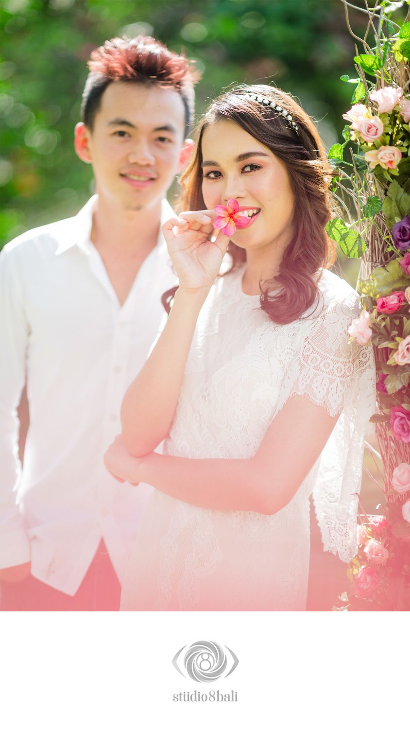 Couple in white outfits pose among pink flowers in a romantic Bali garden engagement photo by Studio 8 Bali.