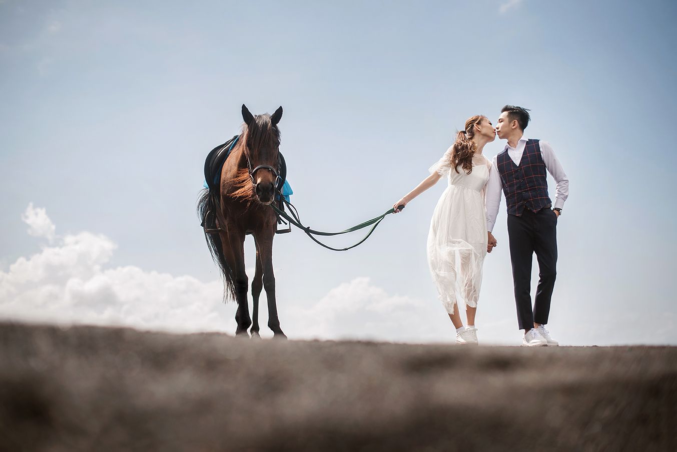 Bride and groom sharing a kiss beside a dark brown horse under a blue sky on their wedding day.