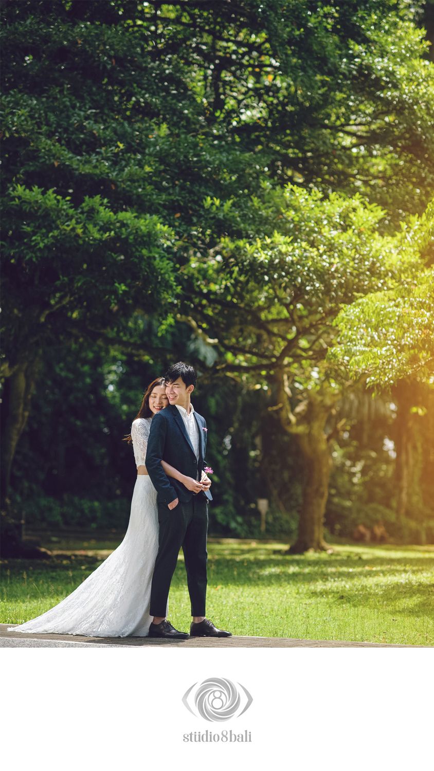 A couple in wedding attire poses romantically among lush green trees in a sunlit park setting.