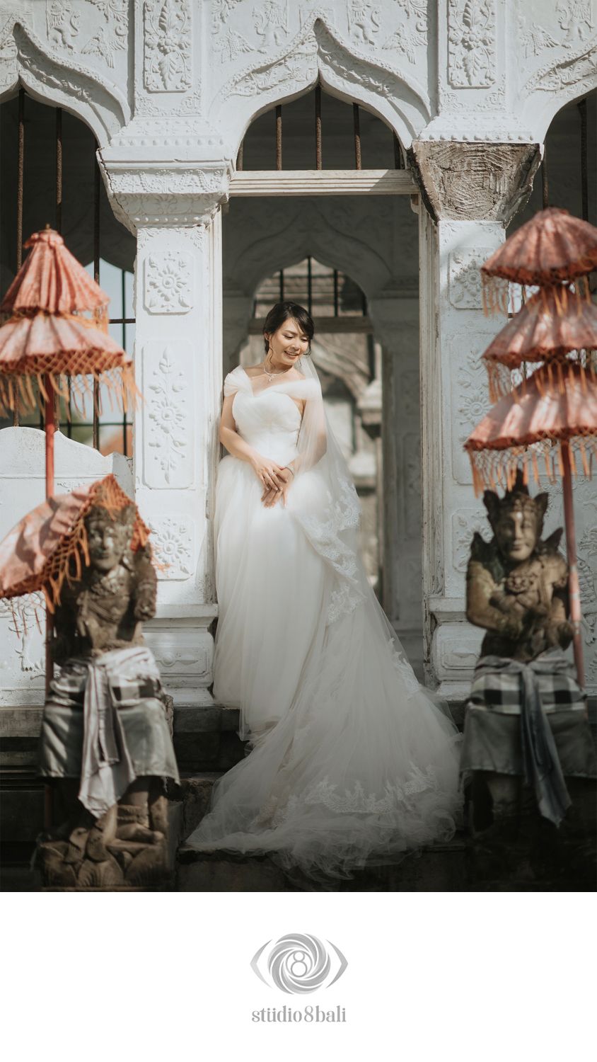 Bride in white wedding gown poses between Balinese stone statues and traditional umbrellas at ornate temple entrance.