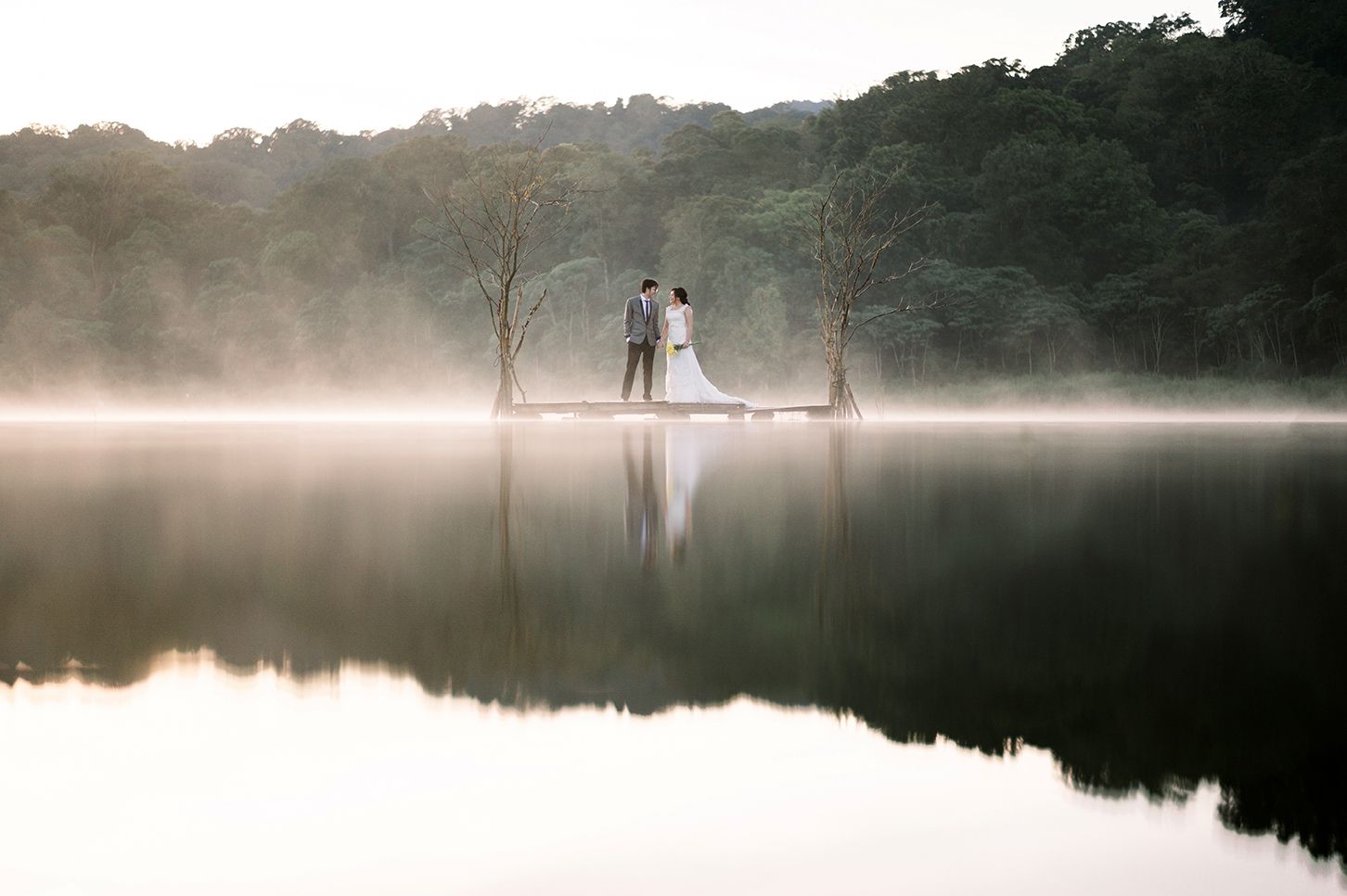 A couple in wedding attire stands on a misty lakeside dock surrounded by fog and lush green trees at dawn.
