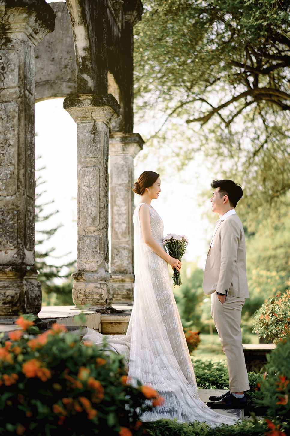Bride in white gown holds bouquet facing groom in beige suit among stone columns and orange flowers in lush garden.