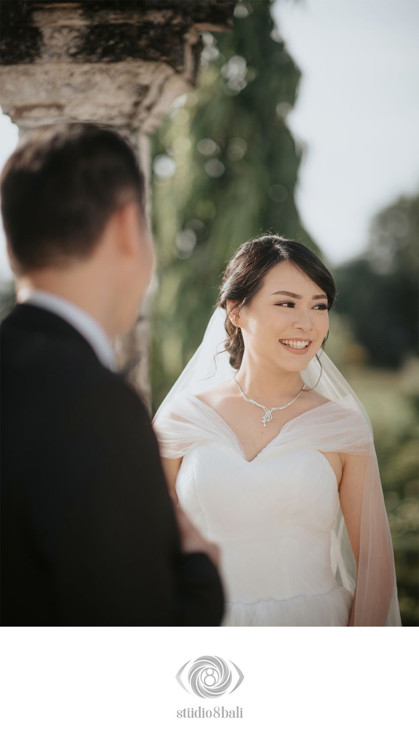 Smiling Asian bride in white wedding dress and veil gazes at groom during outdoor ceremony at historic venue.