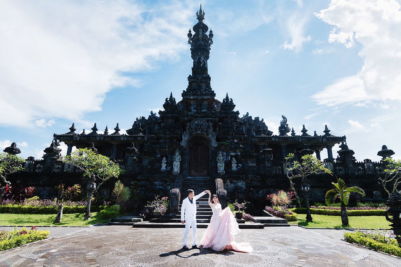 Couple in wedding attire posing before the grand Bajra Sandhi Monument in Bali, Indonesia, under a blue sky.