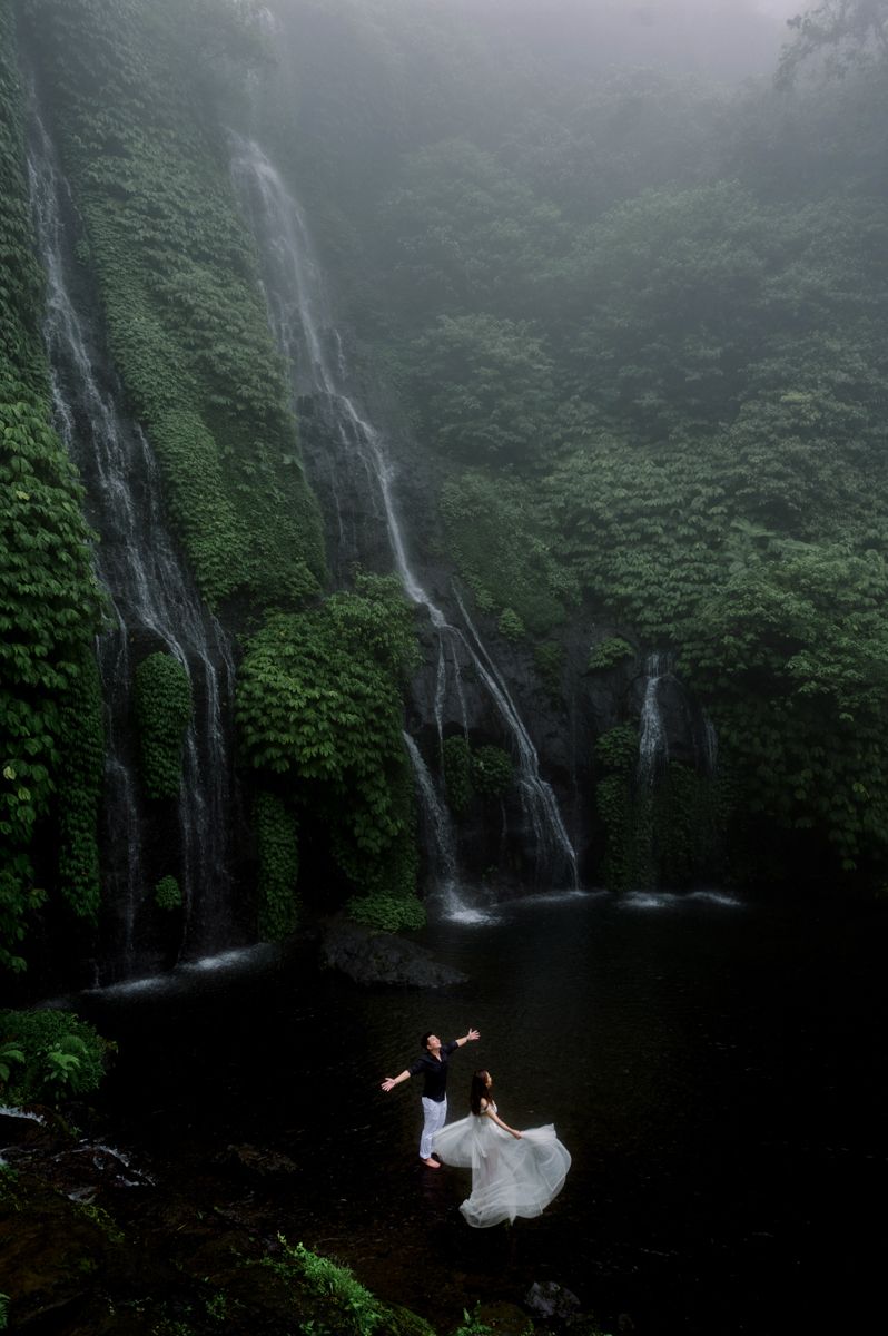 Couple in wedding attire standing before a misty jungle waterfall with lush green cliffs in a romantic outdoor setting.