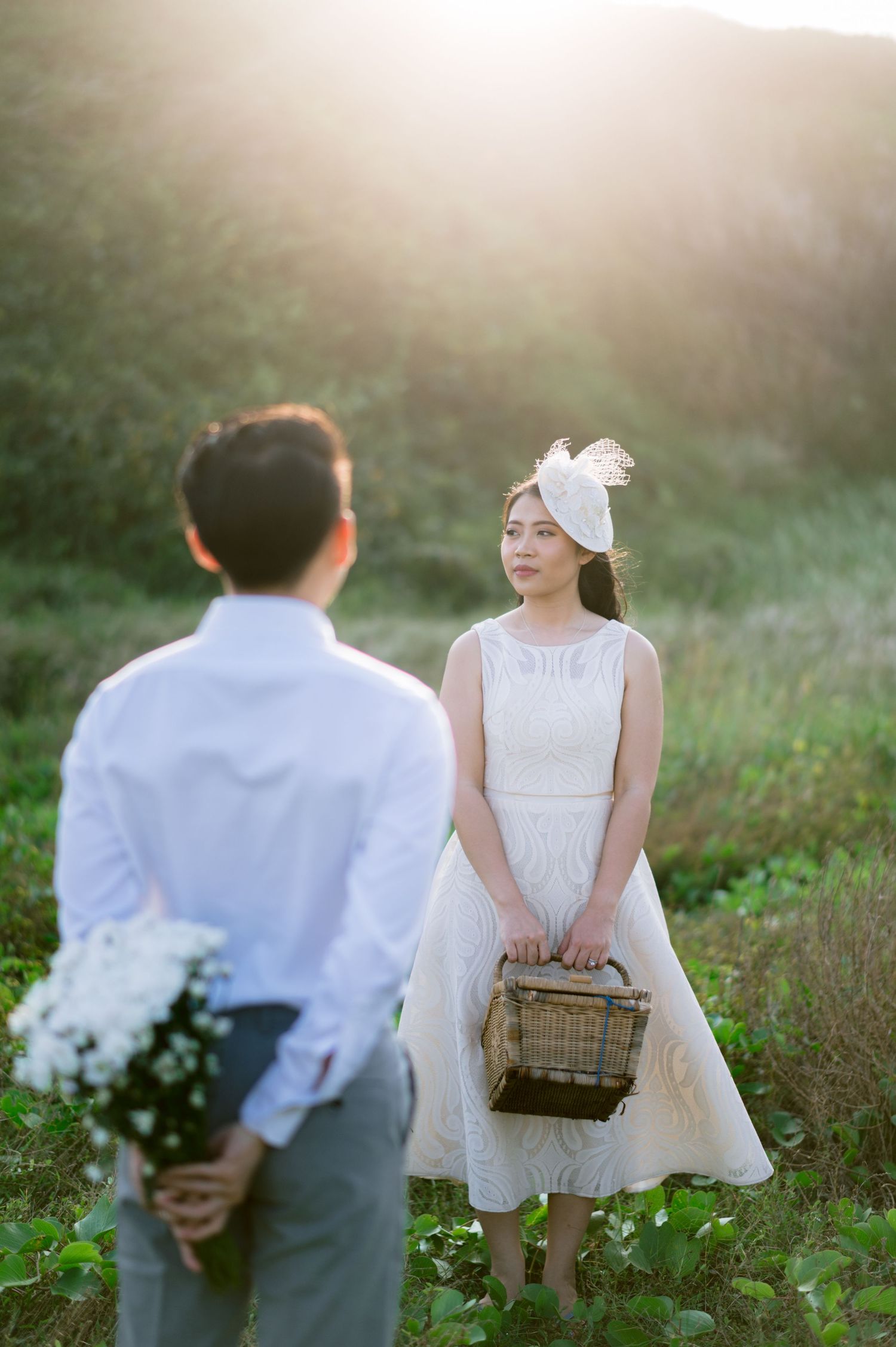 A man holds white flowers behind his back while facing a woman in a white dress and fascinator hat in a sunlit field.