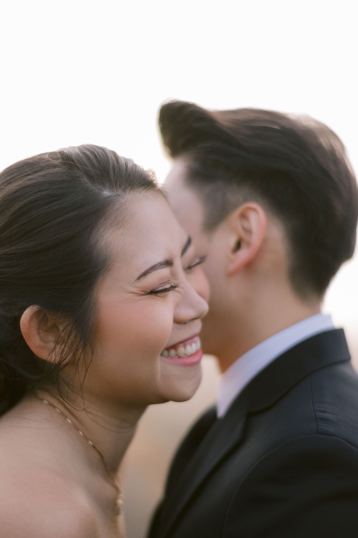 A joyful bride smiles as groom leans in close, captured in a romantic golden-hour wedding portrait.