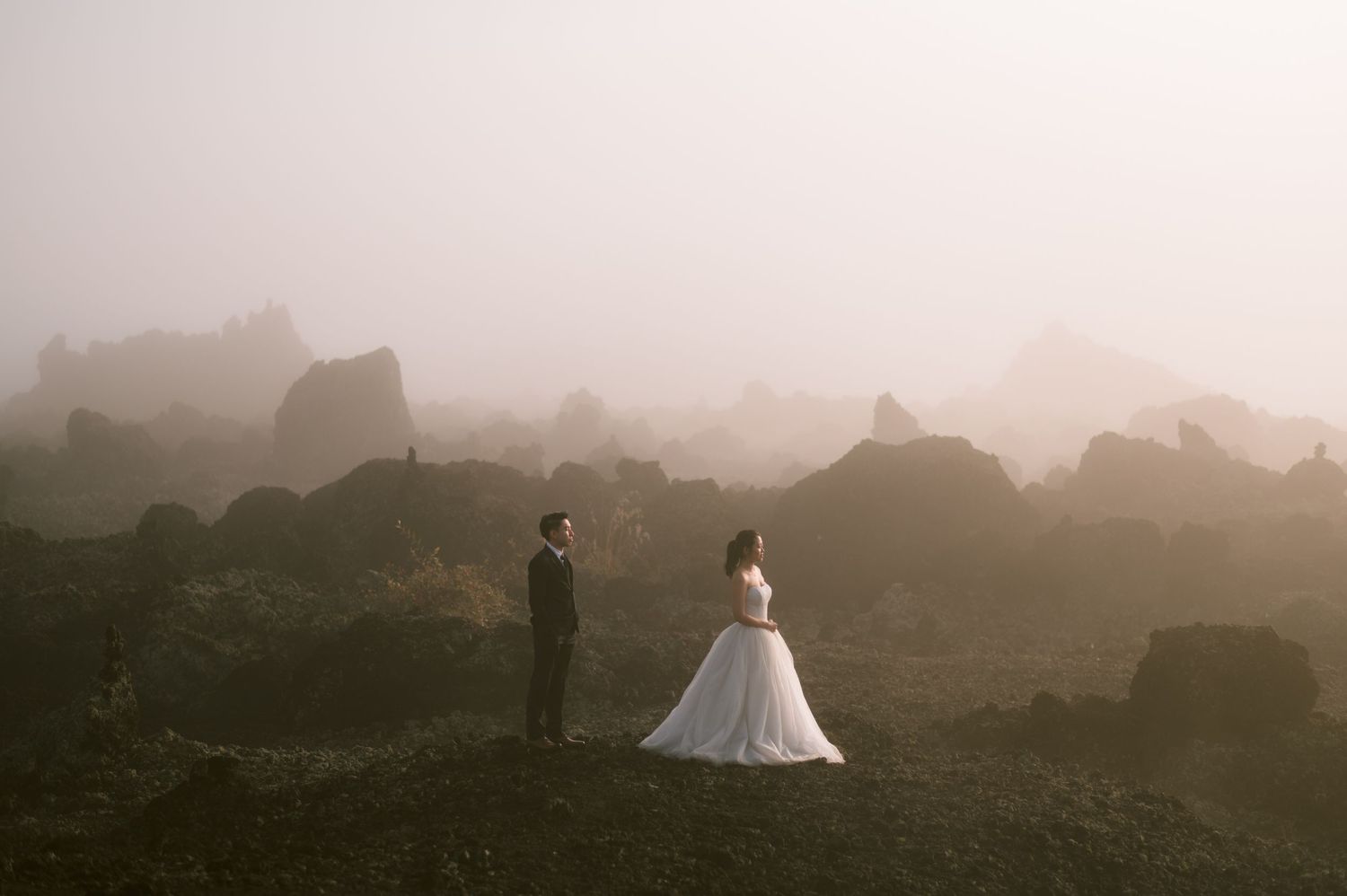 Bride and groom stand apart in misty rocky landscape during ethereal outdoor wedding photoshoot.