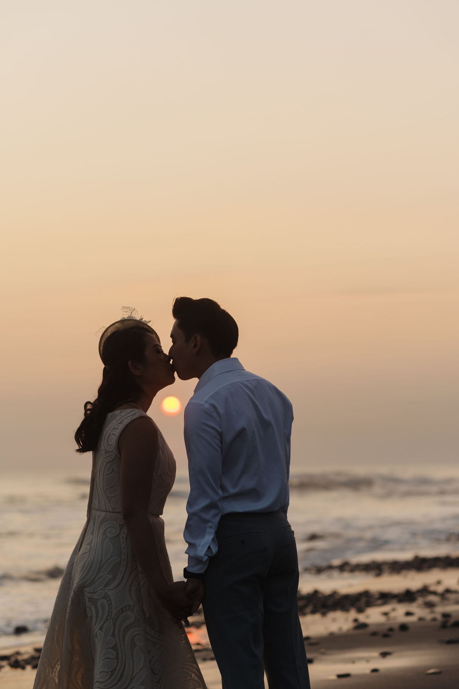 Couple sharing a romantic kiss on the beach at sunset, bride in white dress and groom in blue shirt holding hands.