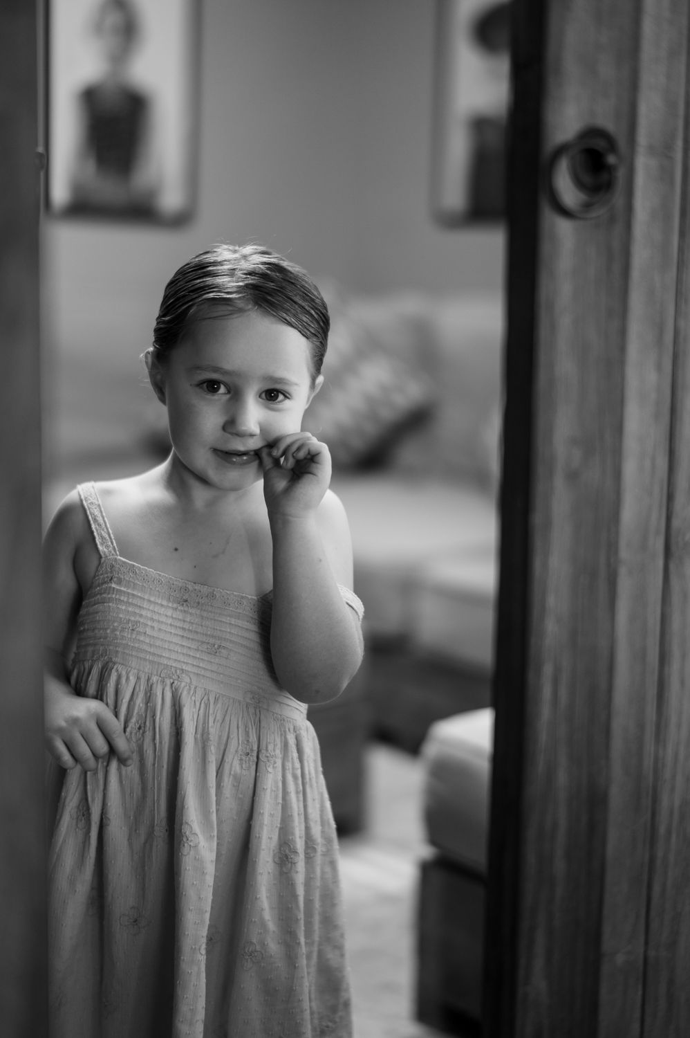 Black and white portrait of a young girl in a sundress standing in a doorway, hand raised to her chin.