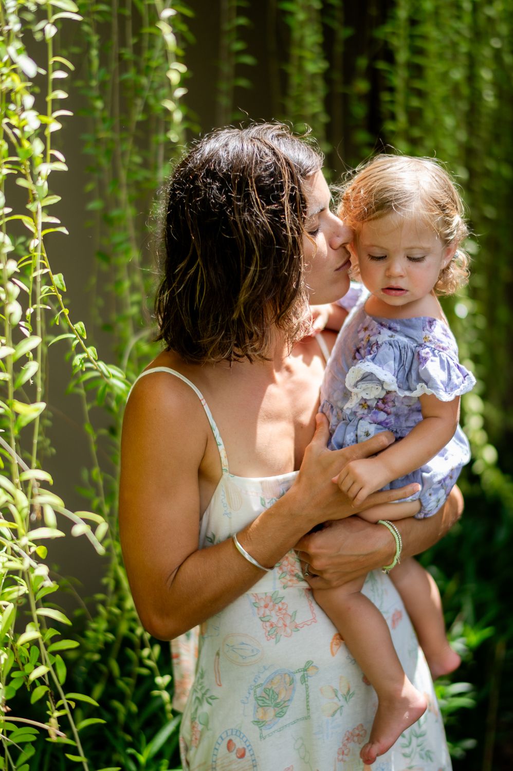 A mother tenderly kisses her toddler daughter outdoors, surrounded by lush green foliage in sunlight.