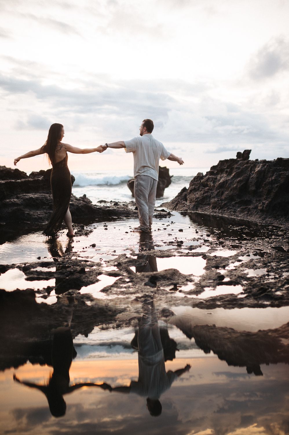 Couple holding hands balancing on rocky coastal tide pools with their reflection in the water at sunset.
