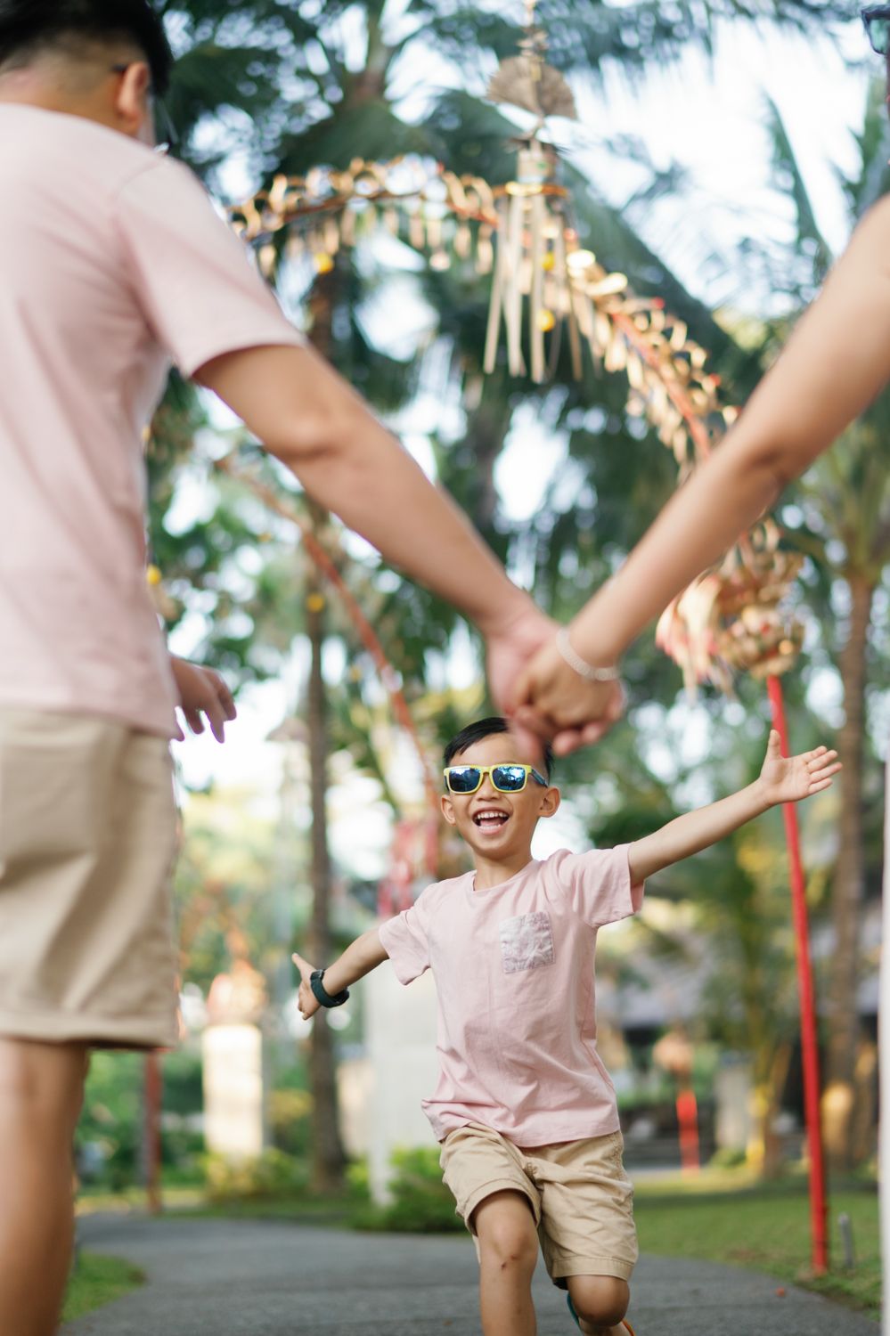 Happy boy in sunglasses runs toward parents holding hands outdoors in a lush tropical park setting.