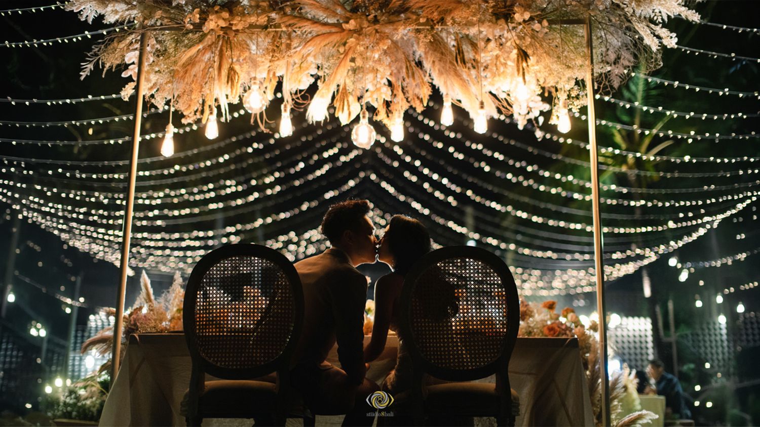 Romantic wedding reception with couple silhouetted under floral canopy and twinkling fairy lights at night.
