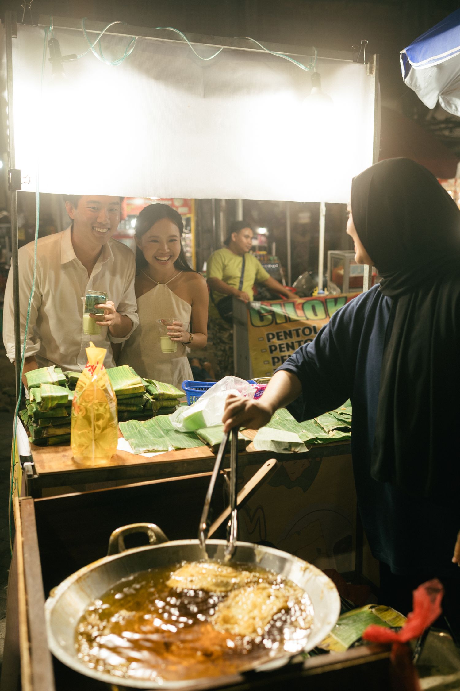 Couple smiling at a night market food stall as vendor cooks in large wok with banana leaves displayed.