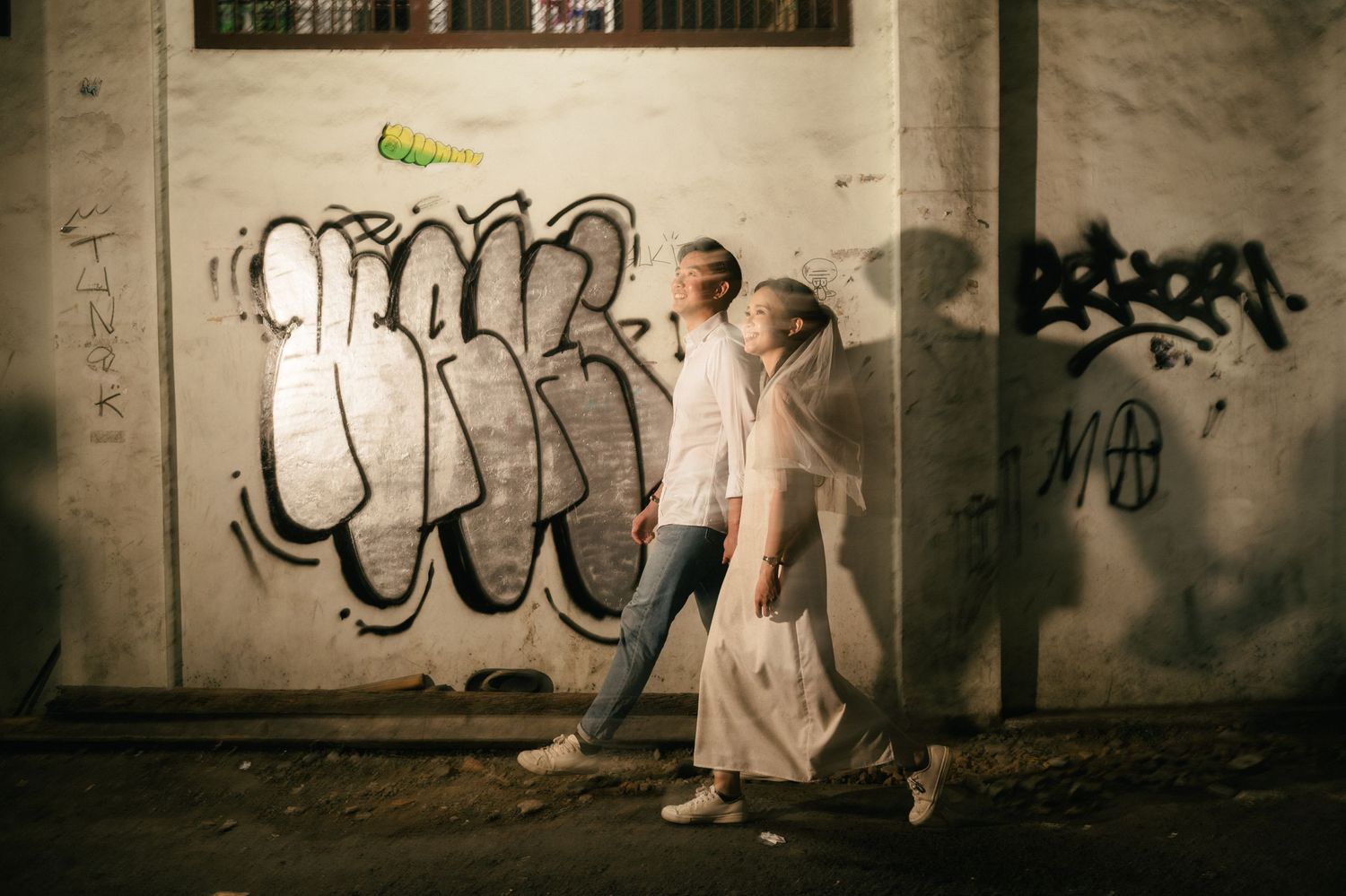 Couple walking past graffiti-covered wall at night in urban street setting.