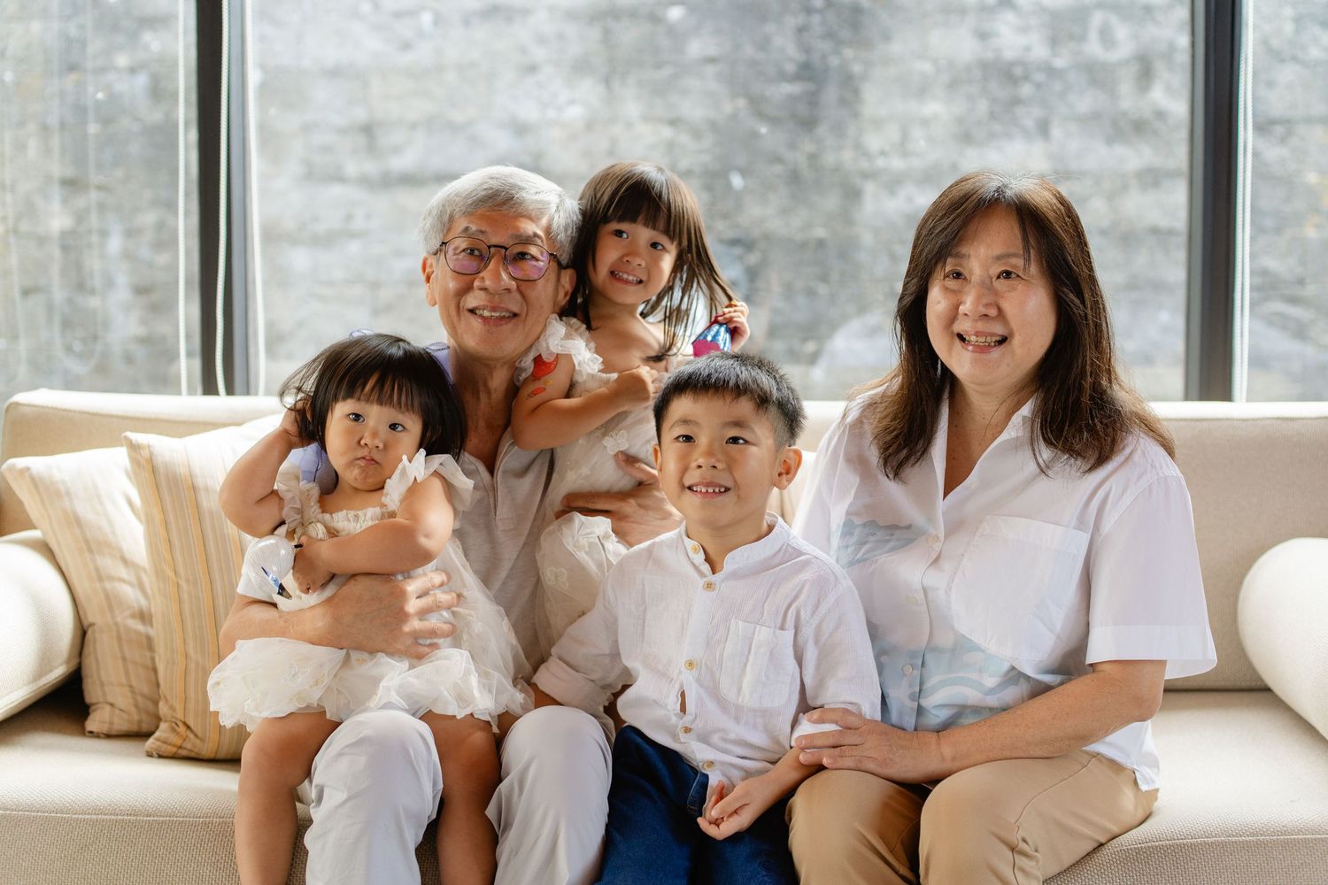 Happy Asian family with grandparents and three young children smiling together on a white sofa near a large window.