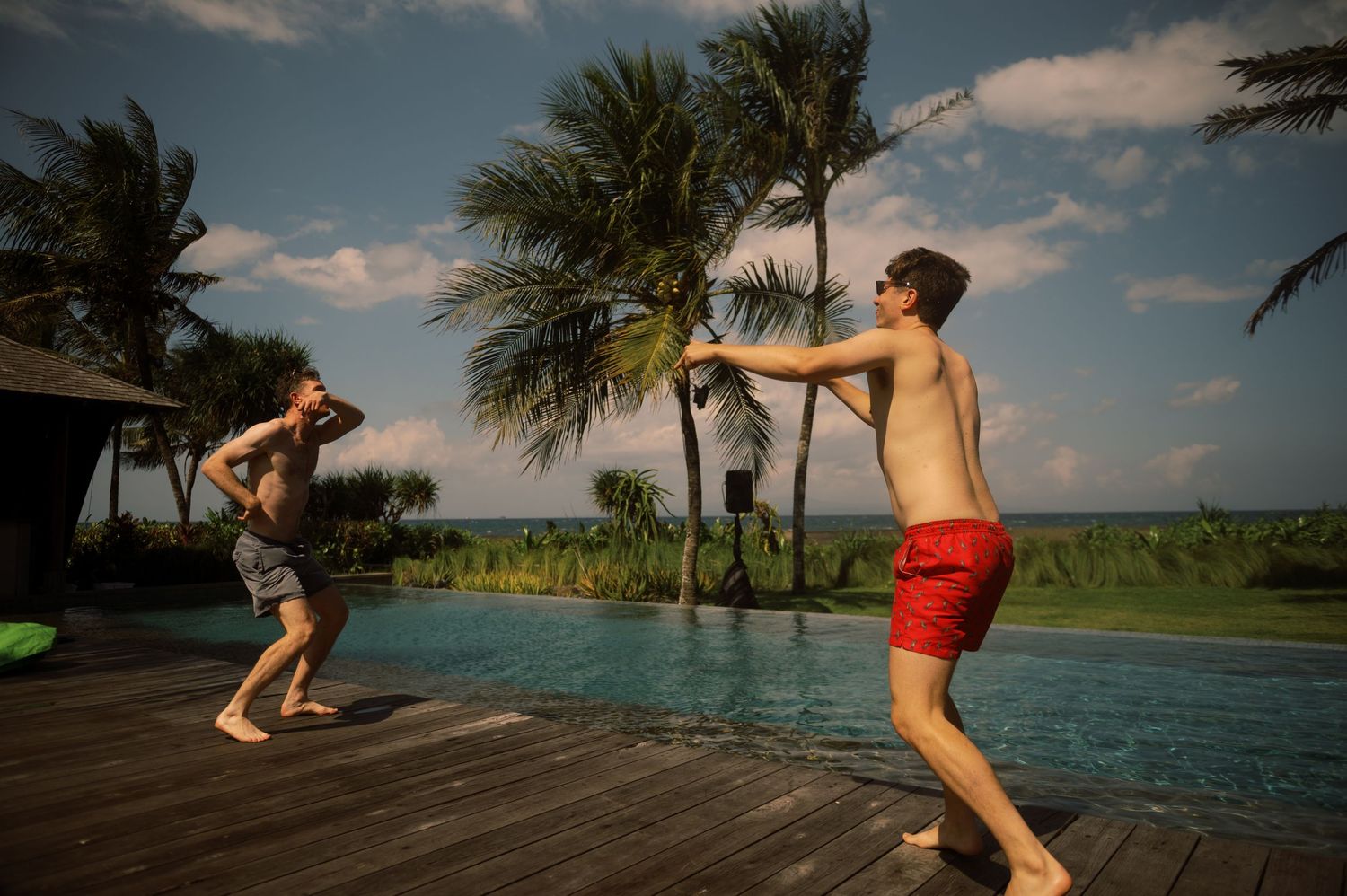 Two shirtless men playing near a tropical infinity pool on a wooden deck with palm trees and ocean views.