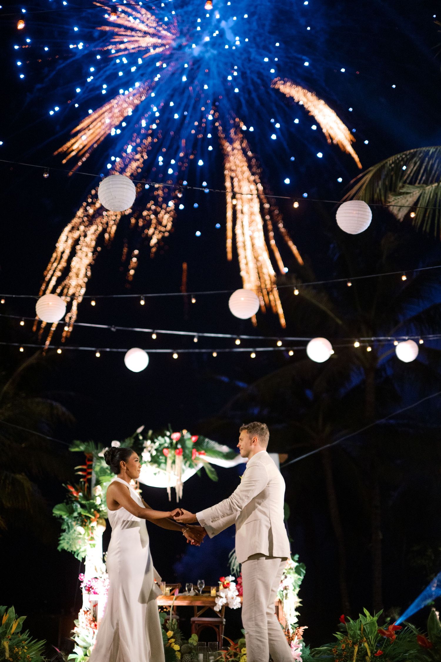 Couple in white attire holds hands at outdoor wedding ceremony with fireworks and string lights at night.