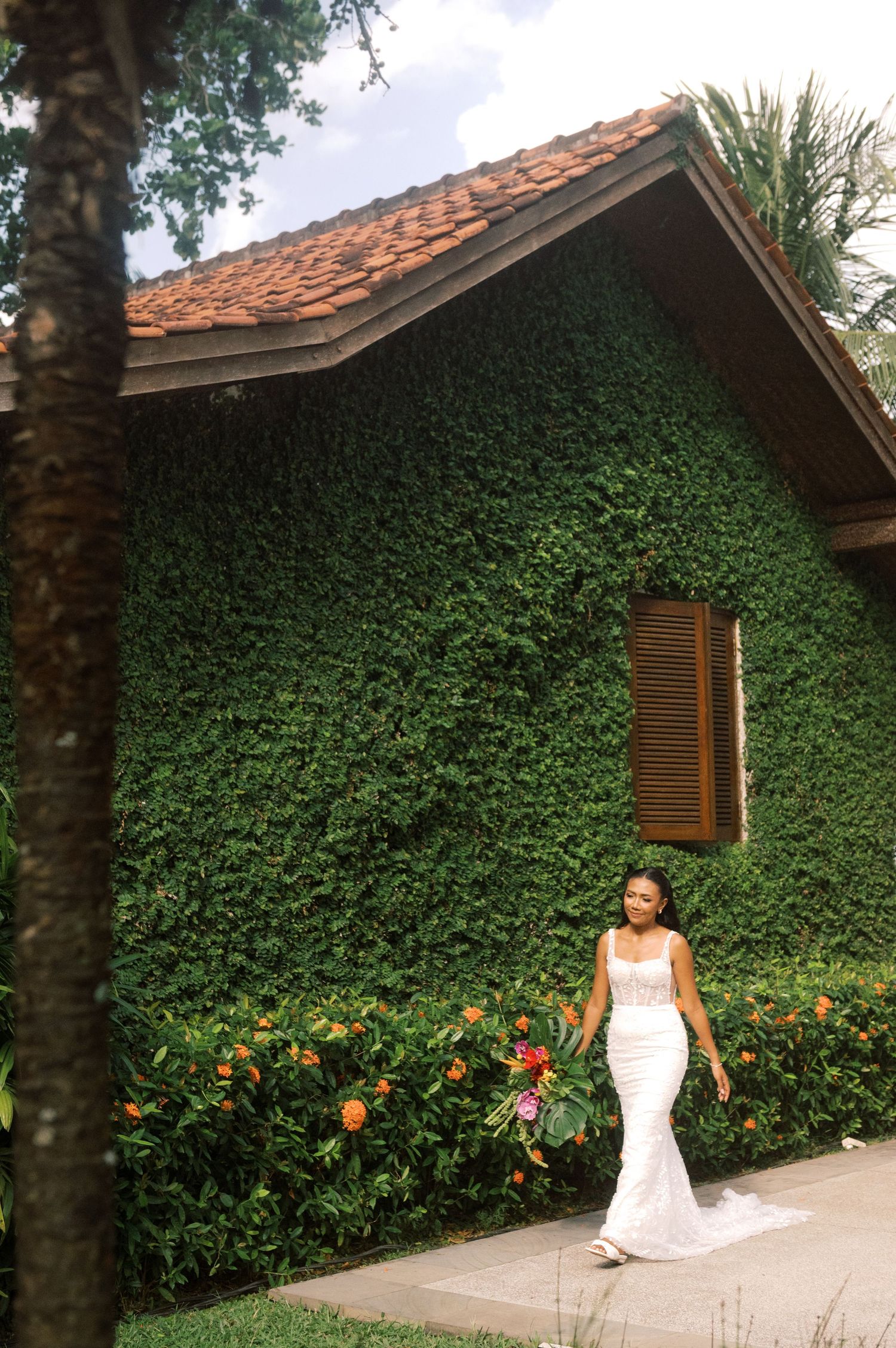 Bride in white gown walks past ivy-covered building with terracotta roof, holding tropical bouquet.