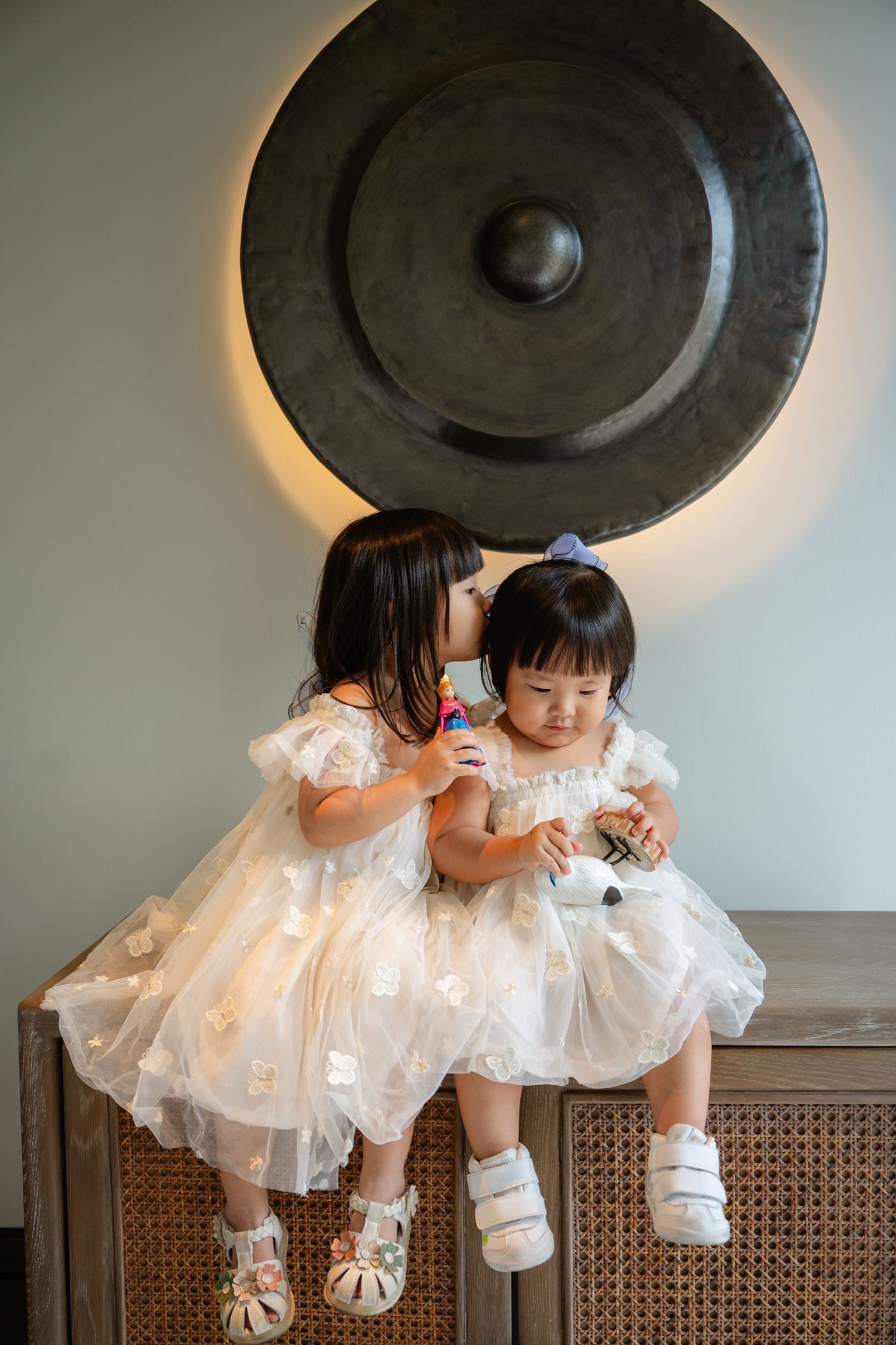 Two young girls in matching white tulle dresses sit together, one kissing the other on the cheek near a decorative gong.