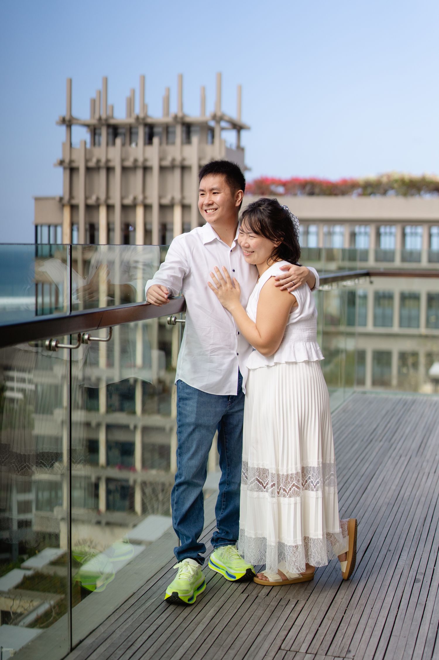 Happy Asian couple embracing on a rooftop balcony, woman showing engagement ring, with urban building in background.