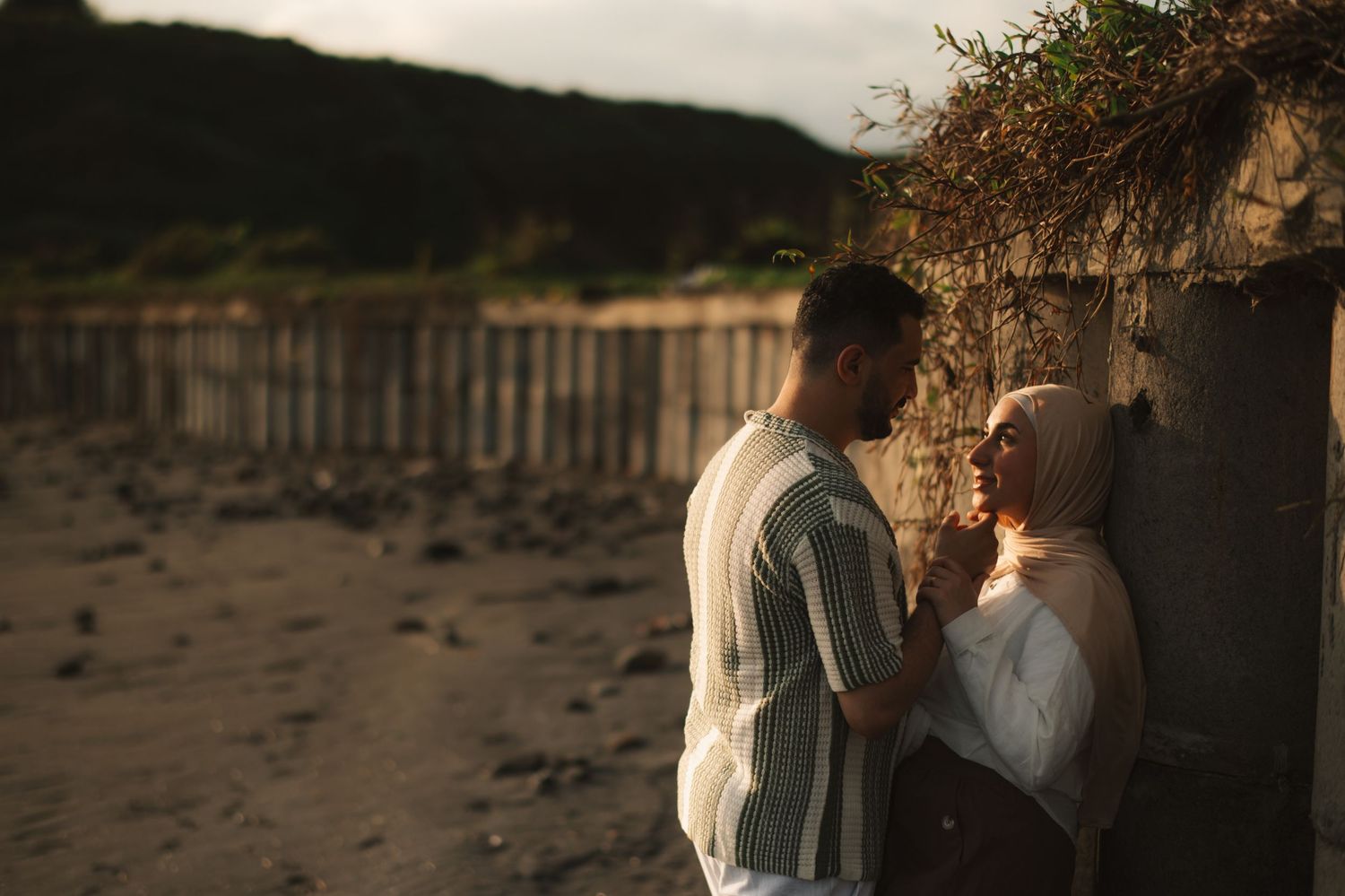 Couple sharing a romantic moment on a sandy beach at sunset near a wooden fence and stone wall.