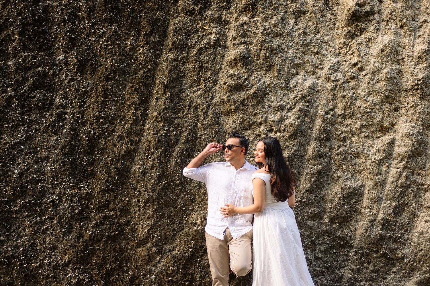 Couple in white outfits posing against a rocky cliff wall, man wearing sunglasses, woman in flowing dress smiling.