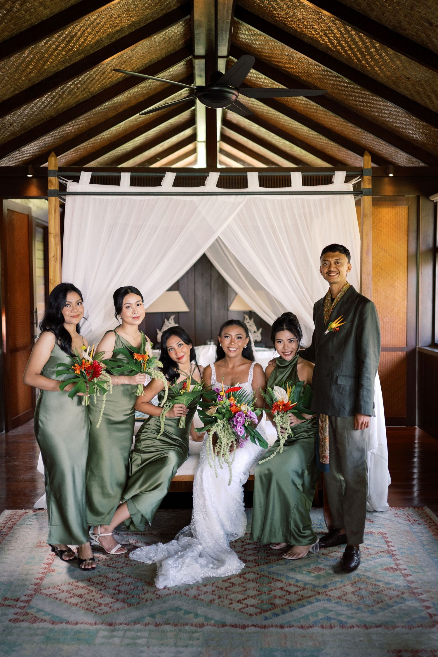 Bride with wedding party in olive green satin gowns pose in tropical villa with thatched ceiling.