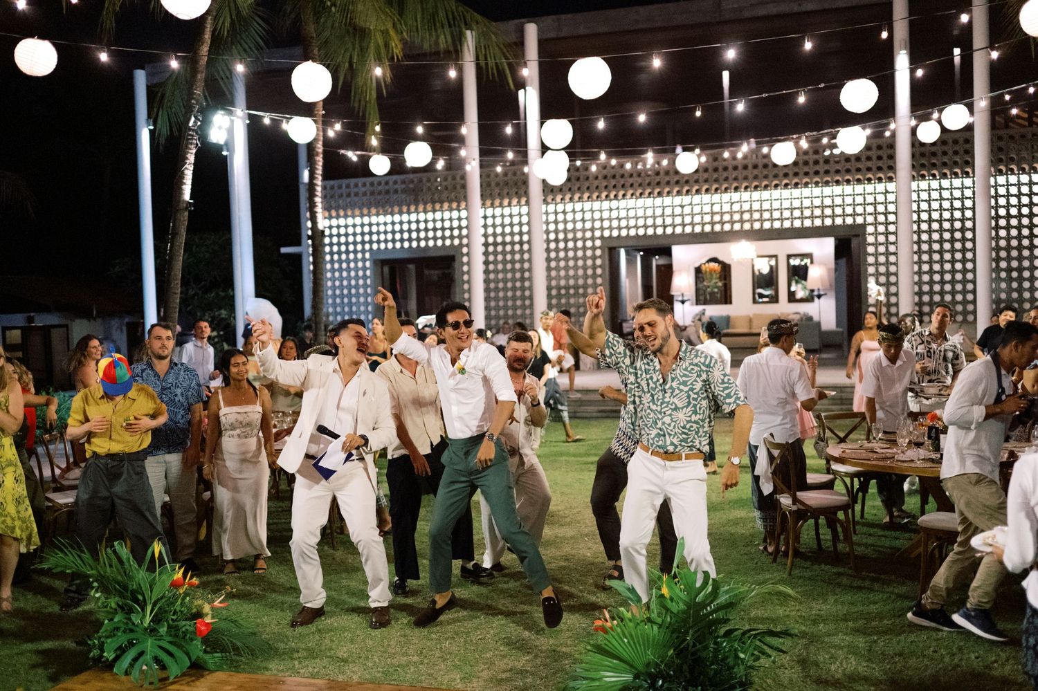 Wedding guests dance energetically on lawn under globe lights at tropical night reception.