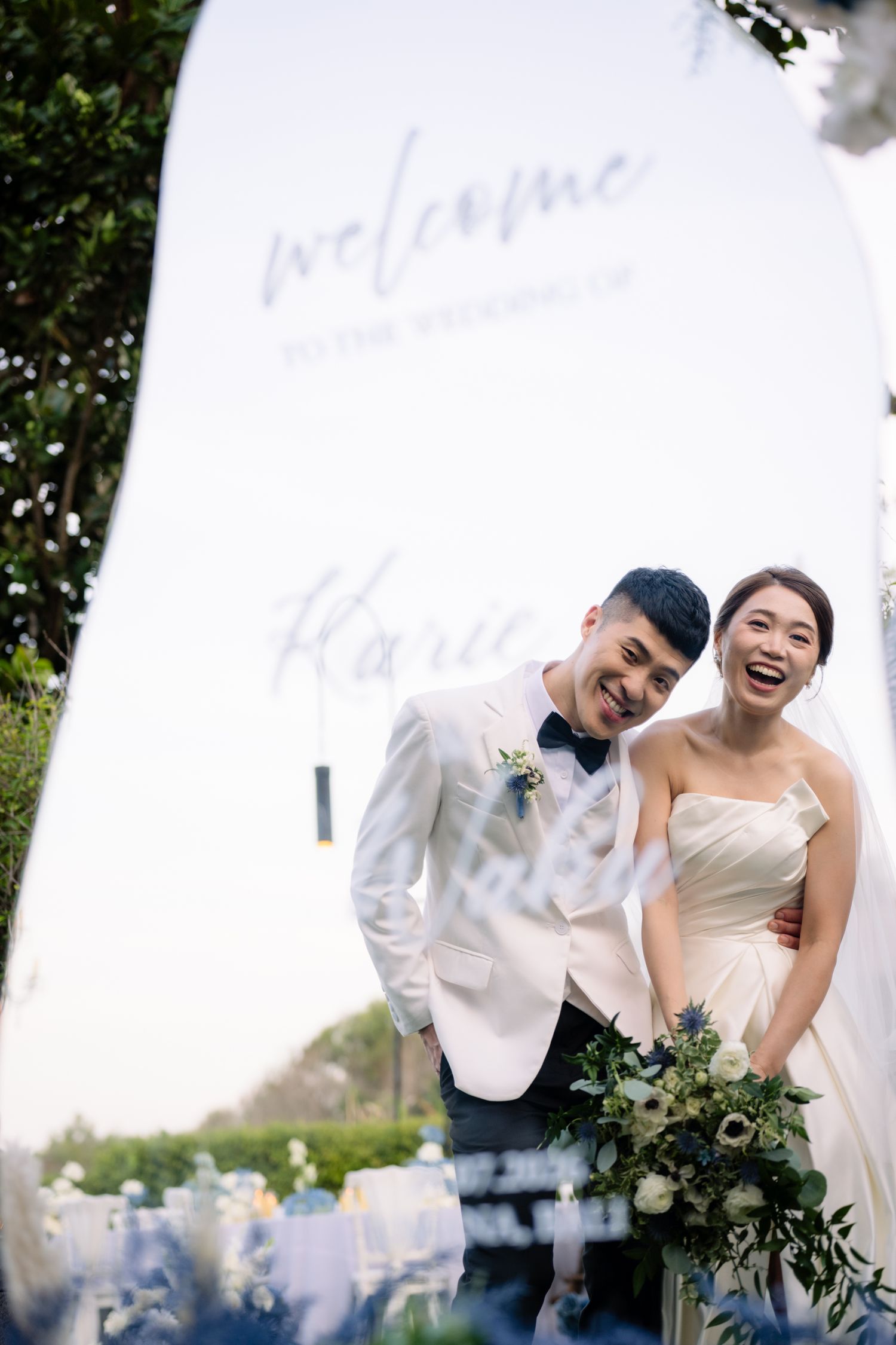 Smiling bride in a strapless white gown and groom in a white tuxedo jacket hold a bouquet beside a welcome to the wedding sign.