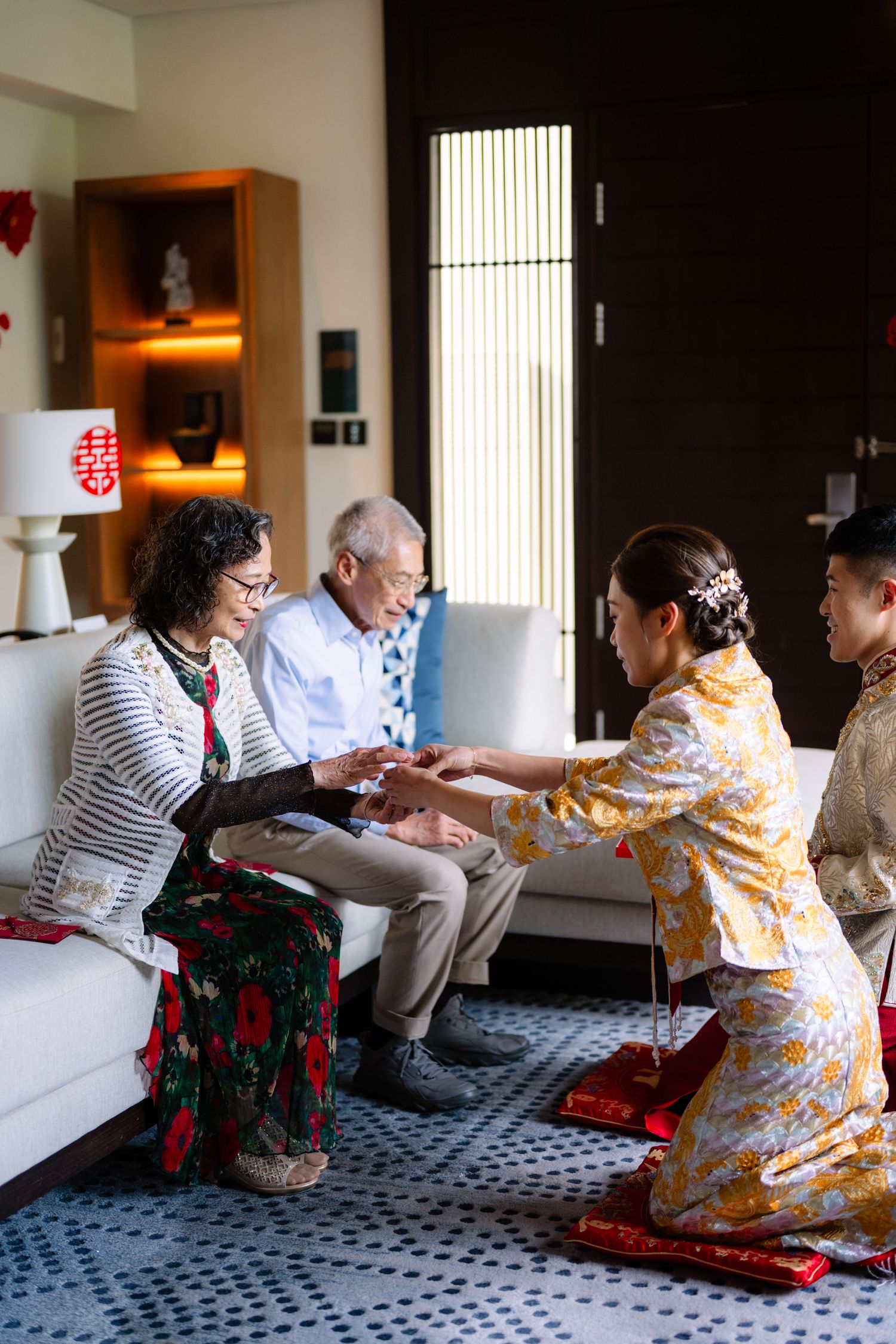 A bride in traditional Chinese wedding attire kneels on a red cushion to serve tea to seated elderly relatives during a tea ceremony.