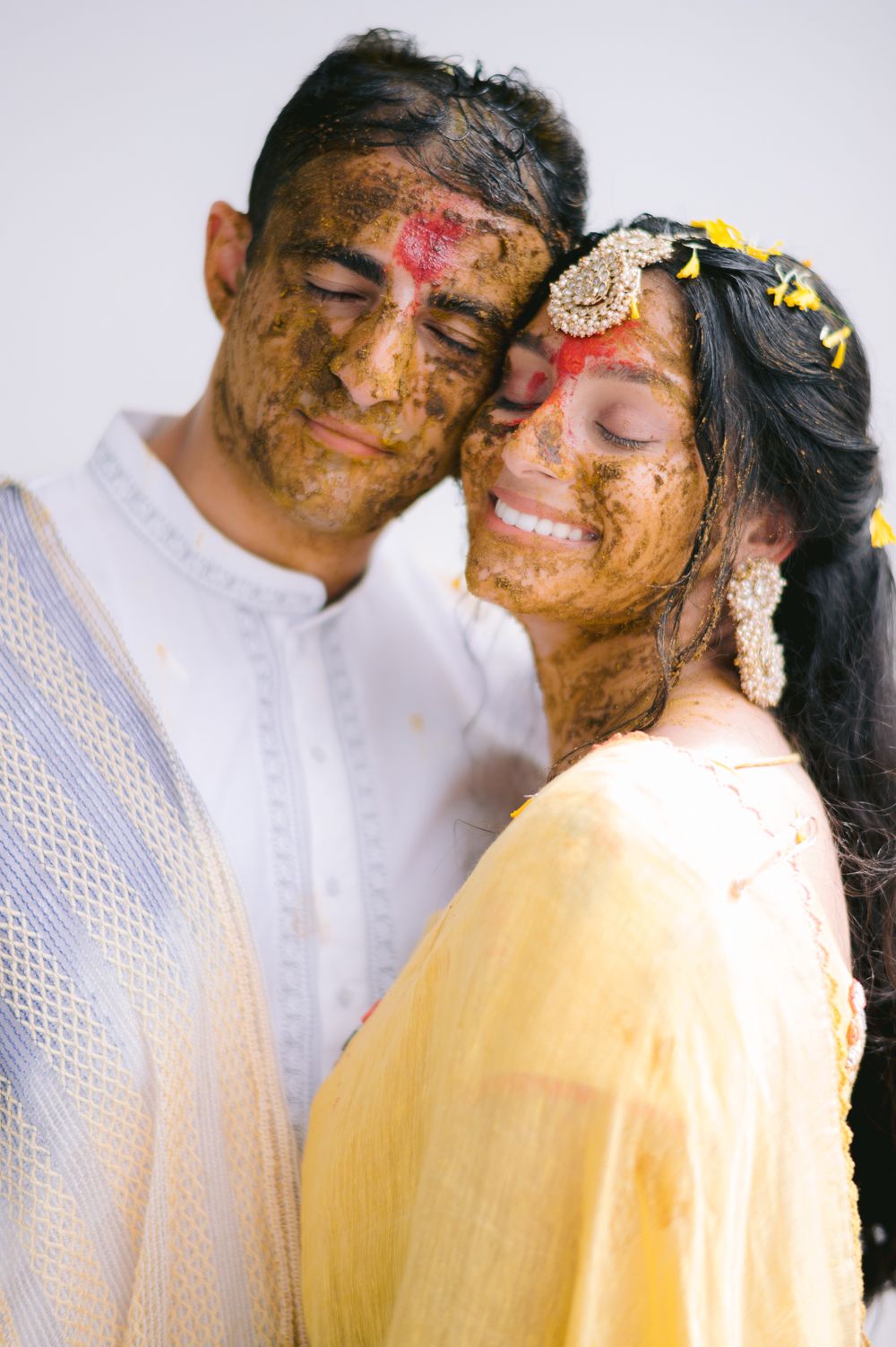 Couple smiling with haldi paste on faces during Indian wedding ceremony, bride in yellow dupatta with floral hair accessories.