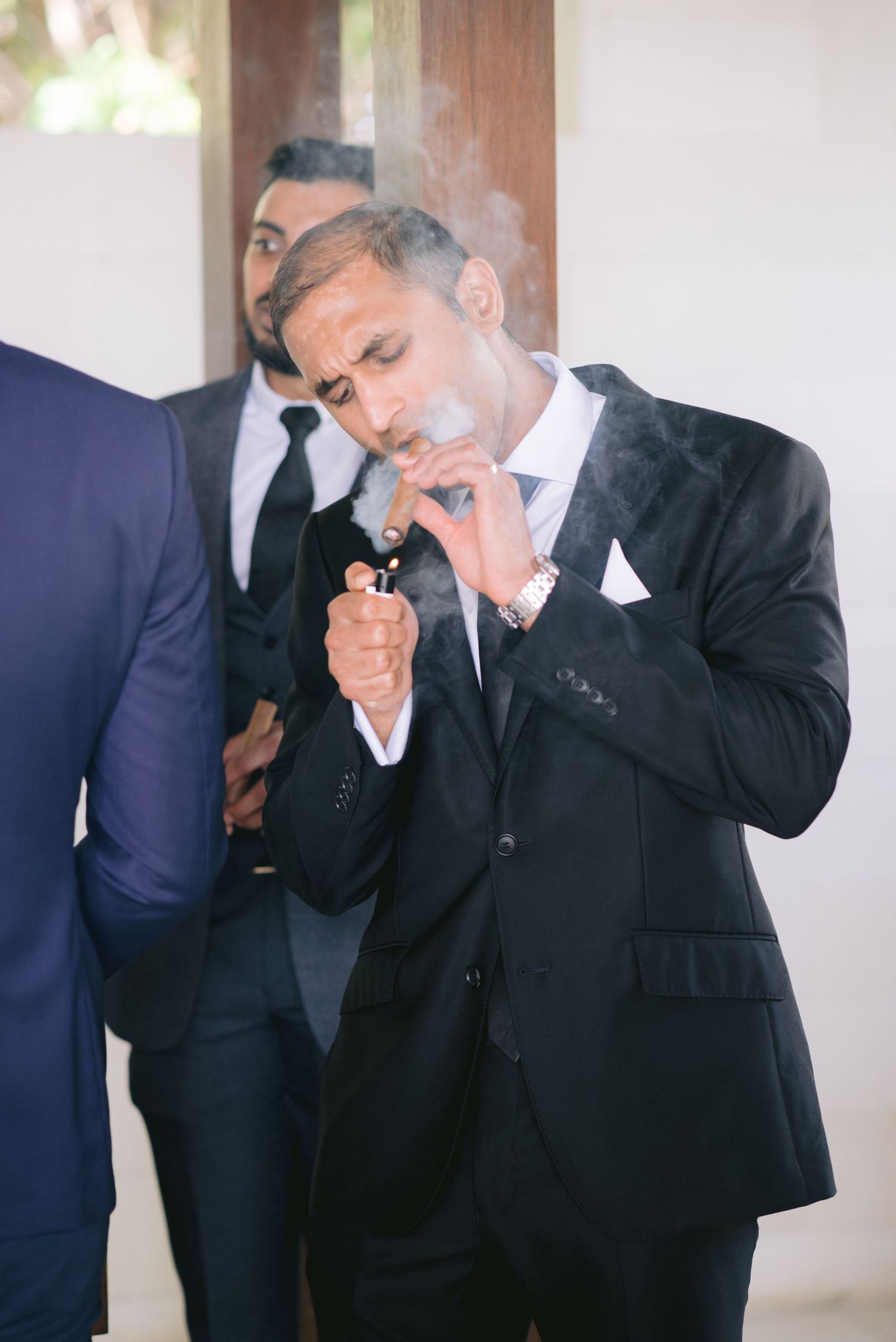A man in a black suit lights a cigar while smoke surrounds him, with another suited man visible behind him.