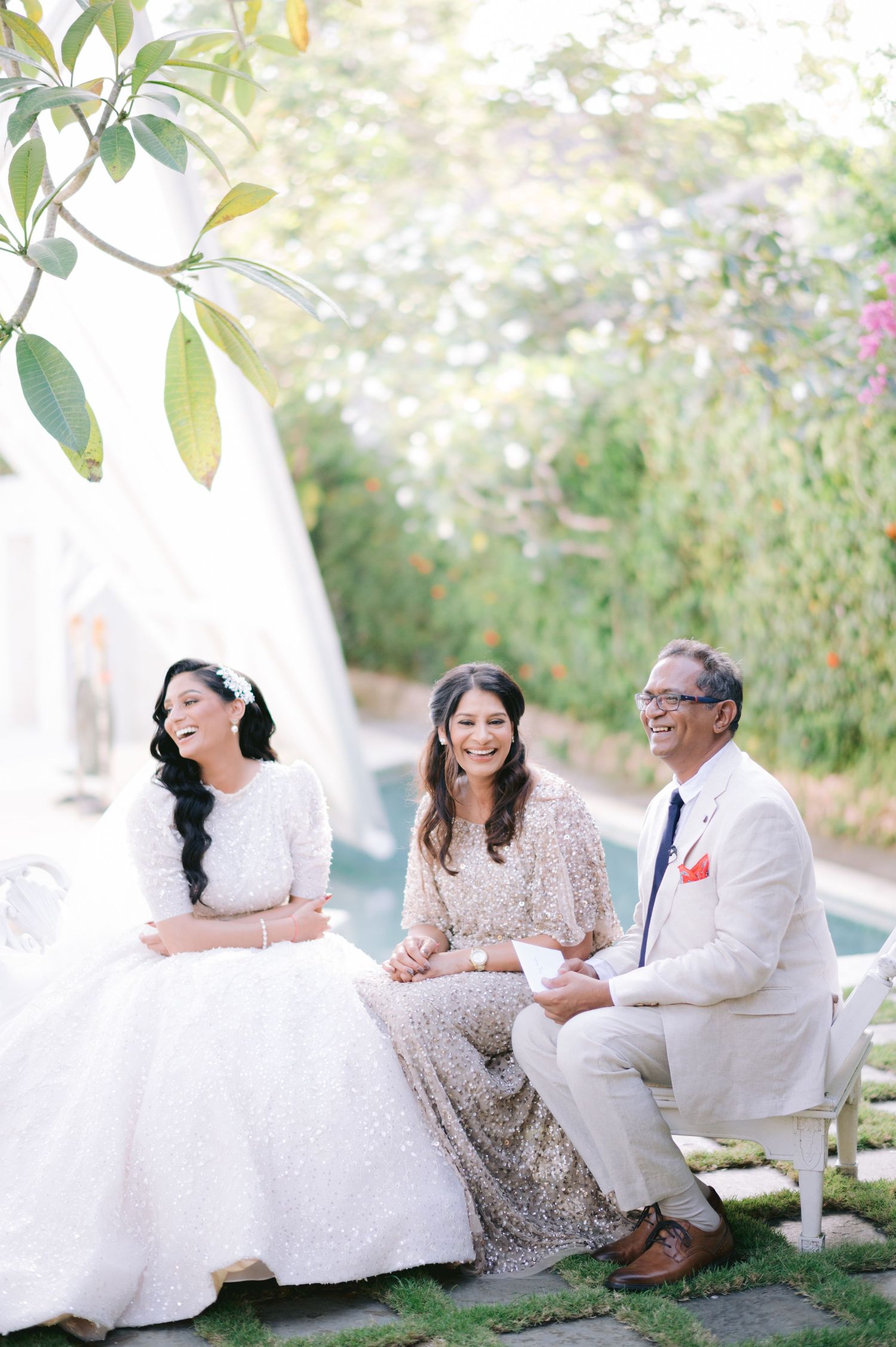 Bride in white gown laughing with family during outdoor wedding ceremony in lush garden setting.