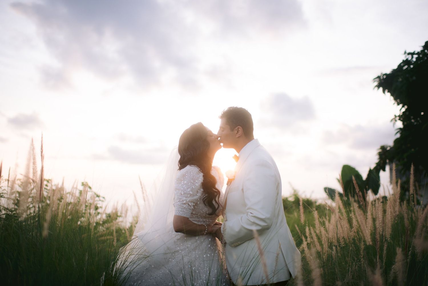 A bride and groom share a romantic kiss at sunset in a lush green field on their wedding day.