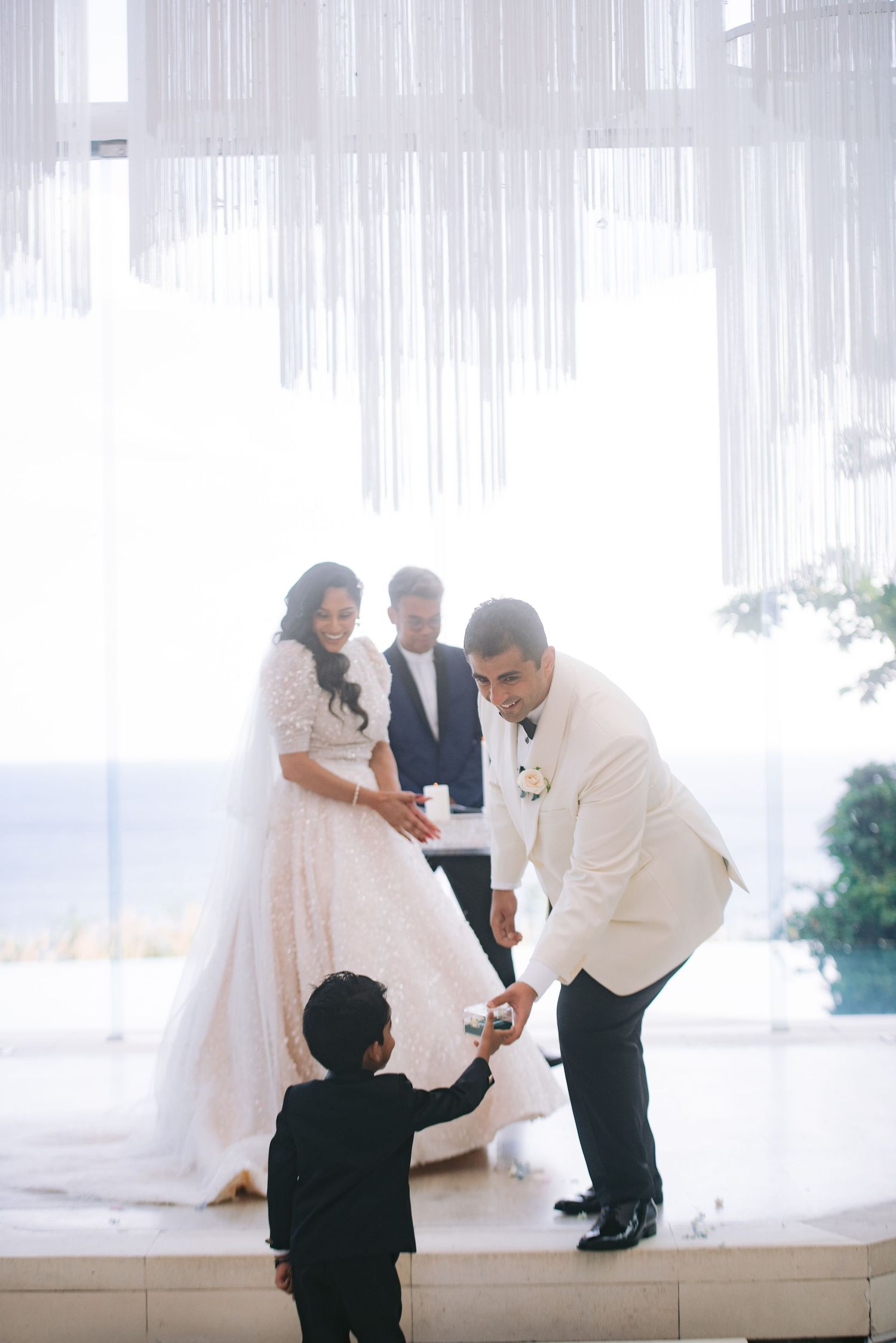 Groom in white tuxedo receives wedding rings from ring bearer during elegant ceremony with bride and officiant.