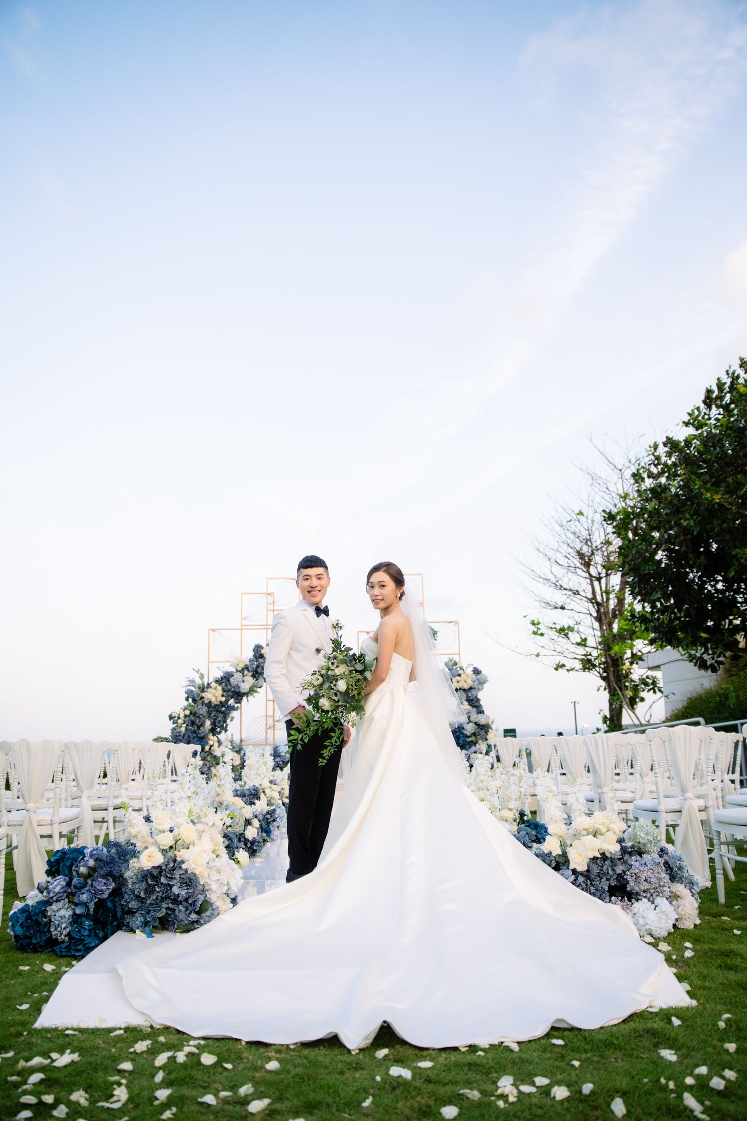 Bride in a flowing white gown and groom in a tuxedo pose on a grassy lawn with blue and white floral arrangements at an outdoor wedding.