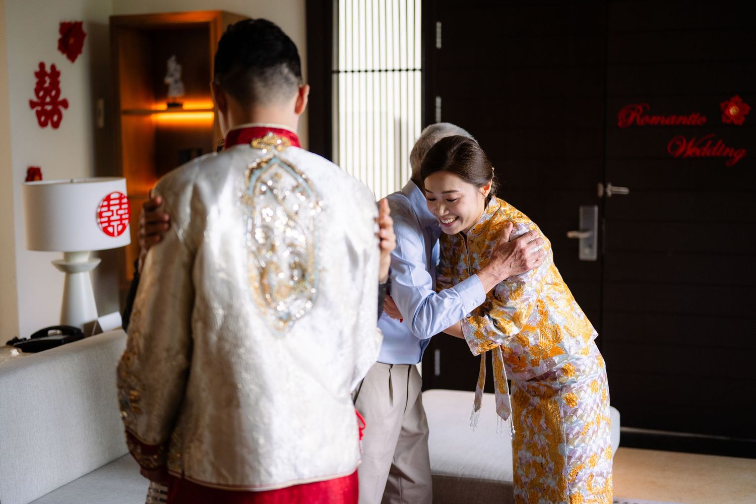 Bride in a gold embroidered Chinese wedding gown embraces a family member while the groom in a traditional ivory jacket looks on.