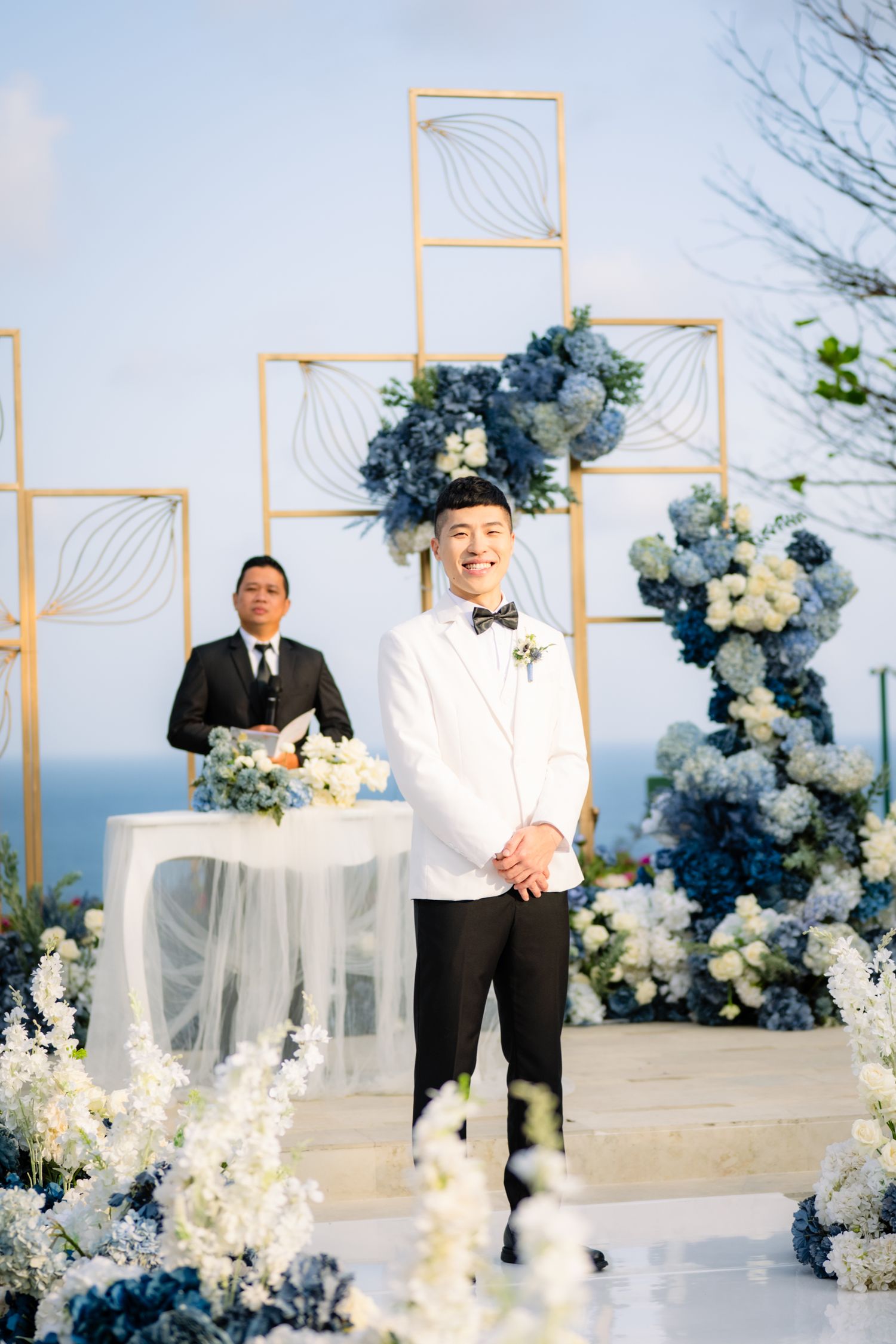 Smiling groom in a white tuxedo jacket and bow tie stands at an oceanfront wedding ceremony with blue and white flowers and gold frames.